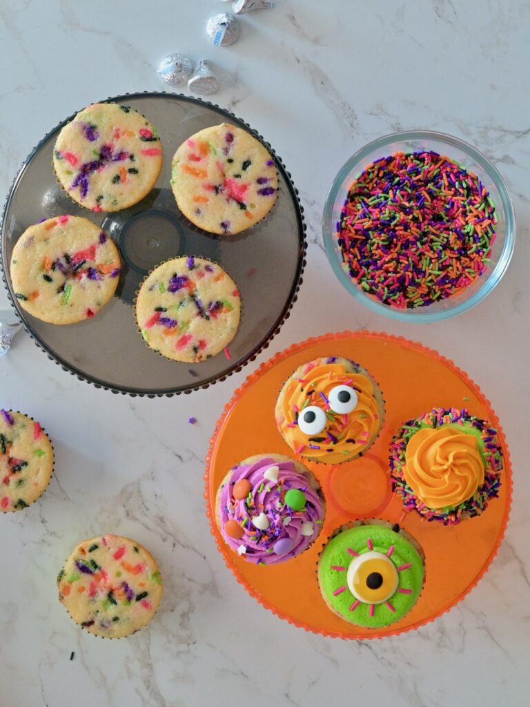 An overhead shot of some baked and unfrosted Halloween confetti cupcakes, and some decorated Halloween cupcakes with orange, purple, and green frosting and topped with Halloween sprinkles and candy eyes.