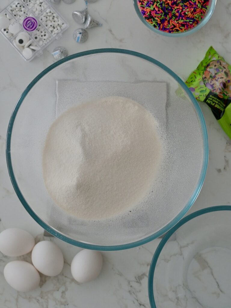 Bowl of sifted dry ingredients for homemade Halloween funfetti cupcakes with candy eyes, sprinkles, and eggs on a marble countertop.