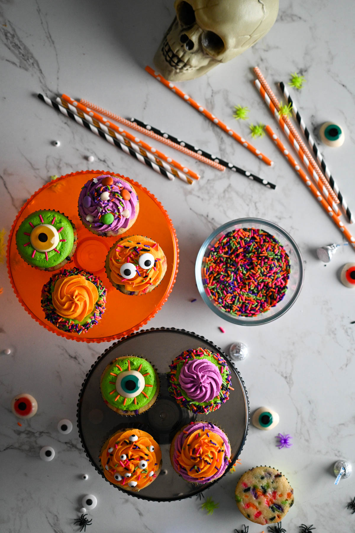 Overhead photo of Halloween cupcakes on orange and black stands surrounded by bowls of rainbow sprinkles, candy eyes, and spooky party decorations.