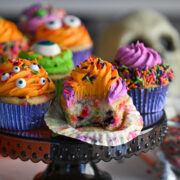 Close-up of colorful Halloween confetti cupcakes in foil cupcake liners, topped with orange, purple, and green frosting, with one open showing the halloween sprinkles inside.