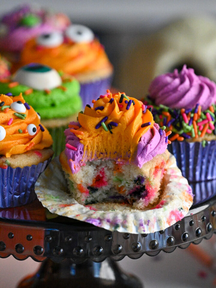Close-up of colorful Halloween confetti cupcakes in foil cupcake liners, topped with orange, purple, and green frosting, with one open showing the halloween sprinkles inside.