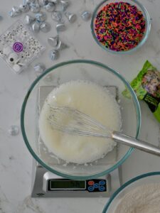 Whisking egg whites in a glass bowl on a kitchen scale surrounded by colorful Halloween sprinkles and candy.