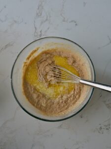 Mixing melted butter into the applesauce cake batter with a whisk in a medium mixing bowl on a marble countertop.