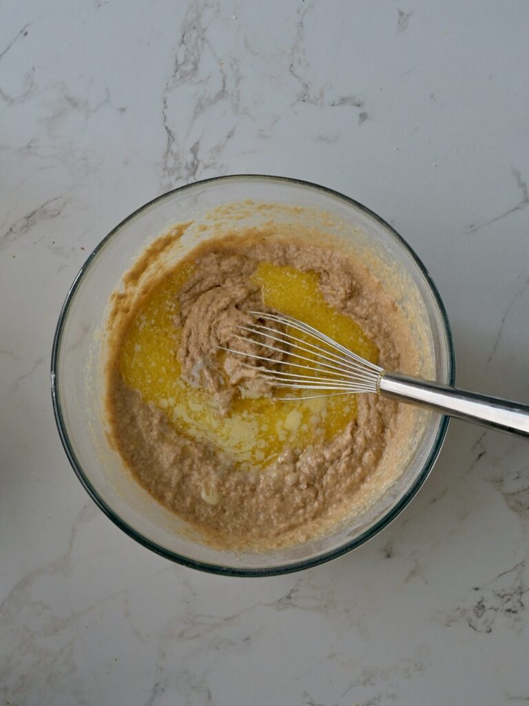 Mixing melted butter into the applesauce cake batter with a whisk in a medium mixing bowl on a marble countertop.