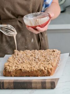 Drizzling cinnamon glaze over a freshly baked applesauce crumb cake on a cutting board, showing the thick crumb layer and soft texture.
