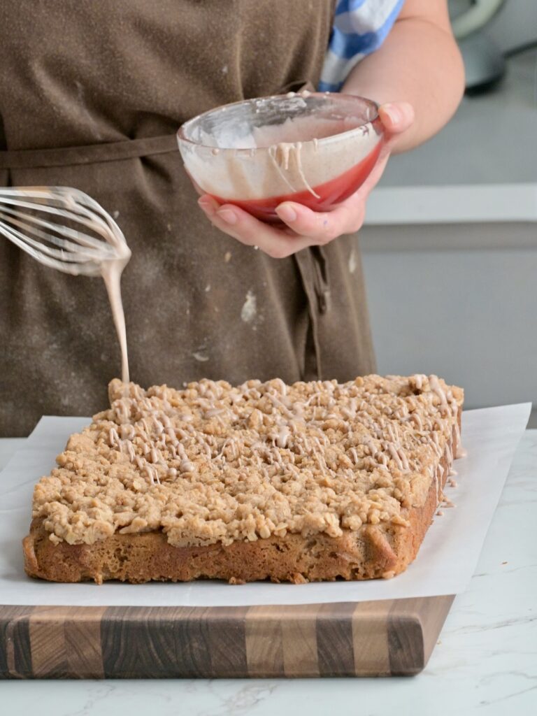 Drizzling cinnamon glaze over a freshly baked applesauce crumb cake on a cutting board, showing the thick crumb layer and soft texture.