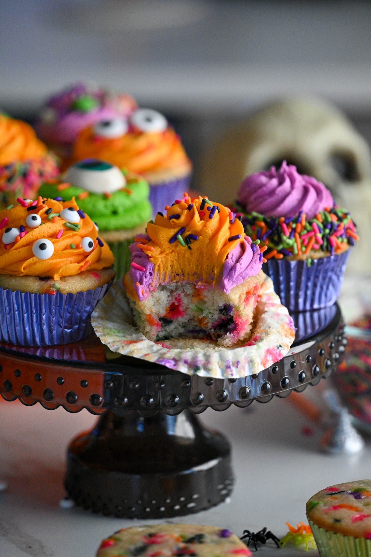 Homemade Halloween Funfetti cupcakes on a black cake stand topped with orange, purple, and green buttercream frosting and colorful Halloween sprinkles, one cupcake cut open to show the confetti center.