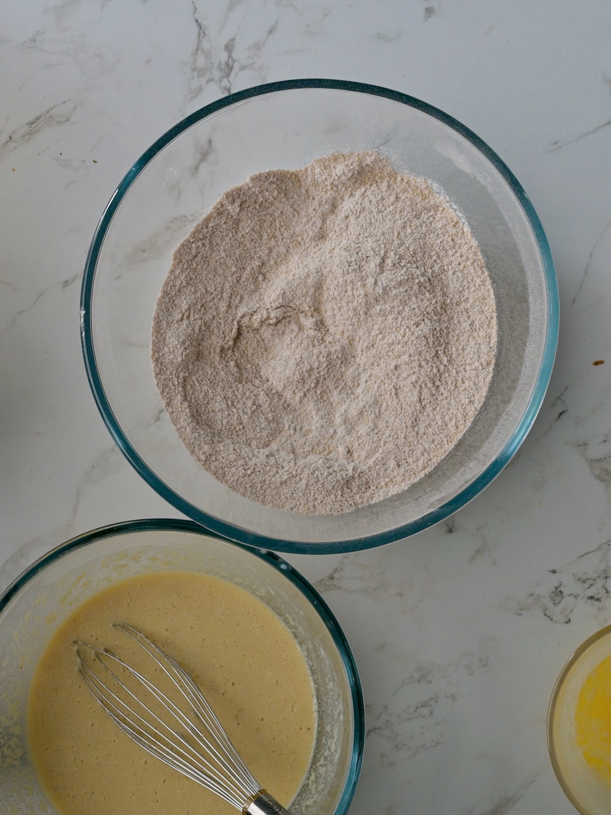 Two mixing bowls side by side showing the wet and dry ingredients for homemade applesauce crumb cake, with a whisk ready for mixing.