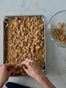 Placing the crumb topping evenly over the applesauce coffee cake batter in a parchment-lined pan before baking.