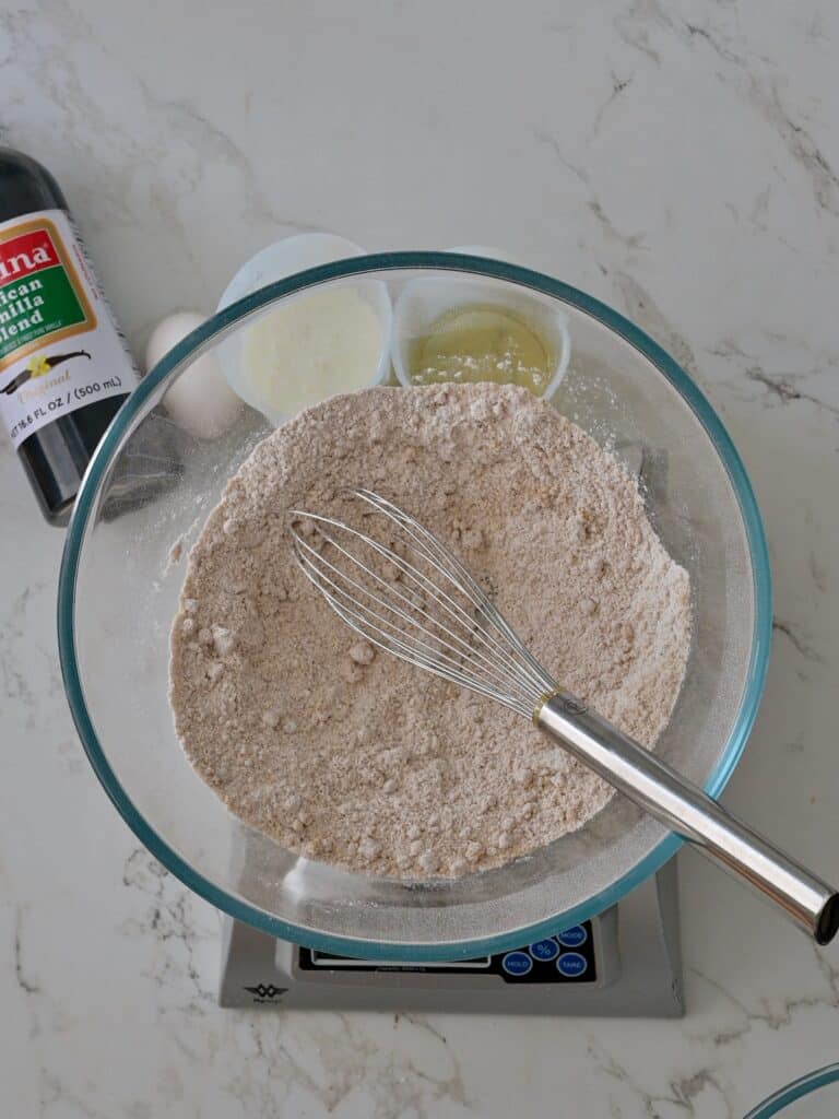 A glass bowl filled with whisked dry ingredients for pumpkin cake, including flour, spices, and brown sugar, sitting on a kitchen scale with vanilla extract and eggs nearby.