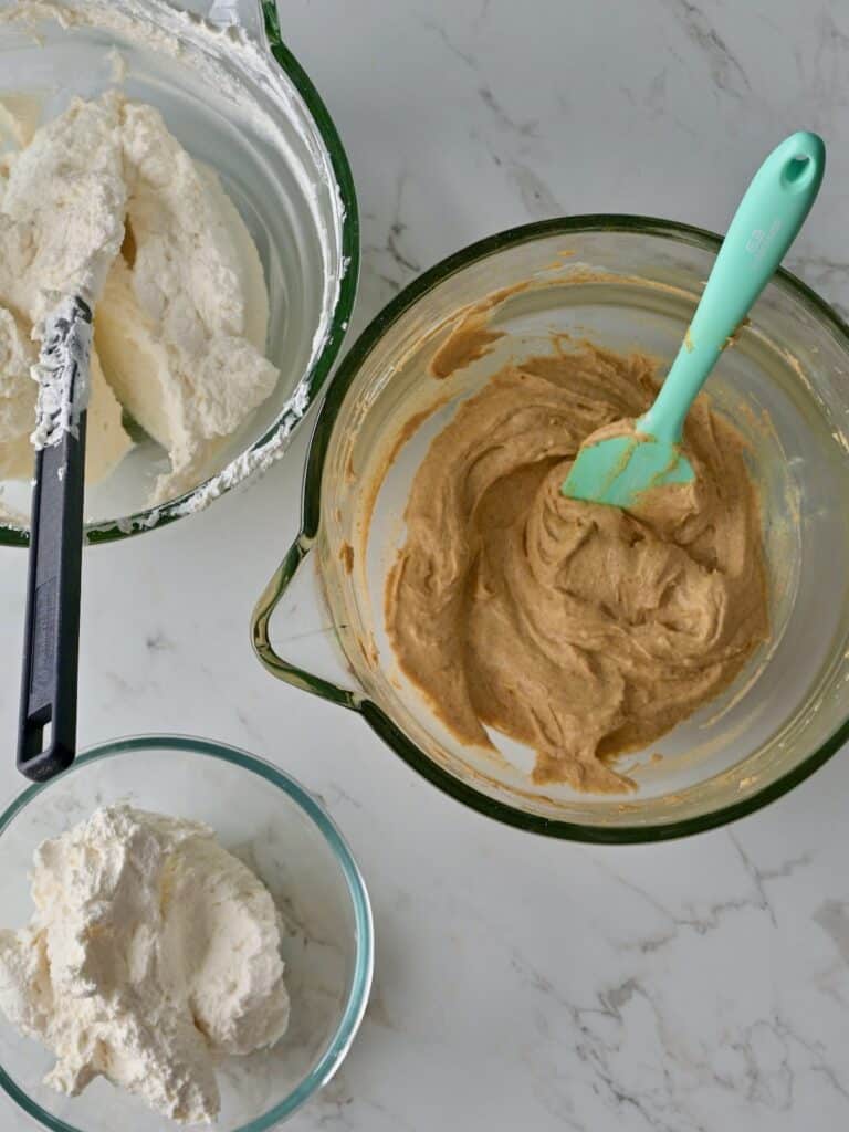 Two bowls of whipped cream next to a bowl of pumpkin mousse base, showing the reserved portions for mousse and frosting the pumpkin cake cheesecake.