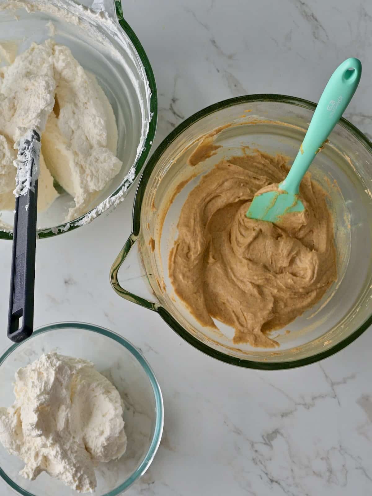 Two bowls of whipped cream next to a bowl of pumpkin mousse base, showing the reserved portions for mousse and frosting the pumpkin cake cheesecake.