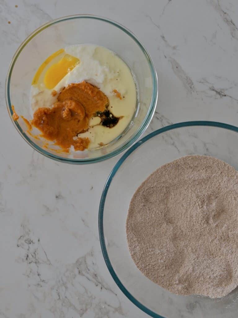 A bowl of wet pumpkin cake ingredients (pumpkin purée, egg, buttermilk, and vanilla) ready to be whisked together beside a second bowl of dry ingredients.