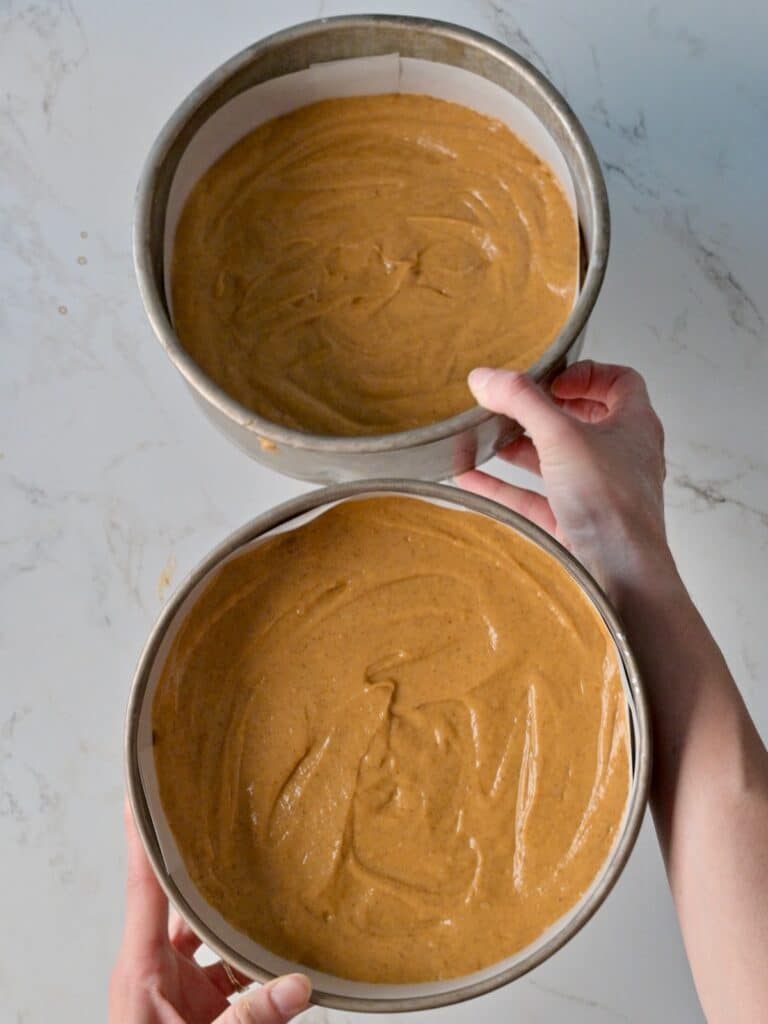 Two parchment-lined 8-inch cake pans filled evenly with pumpkin cake batter, ready to bake.