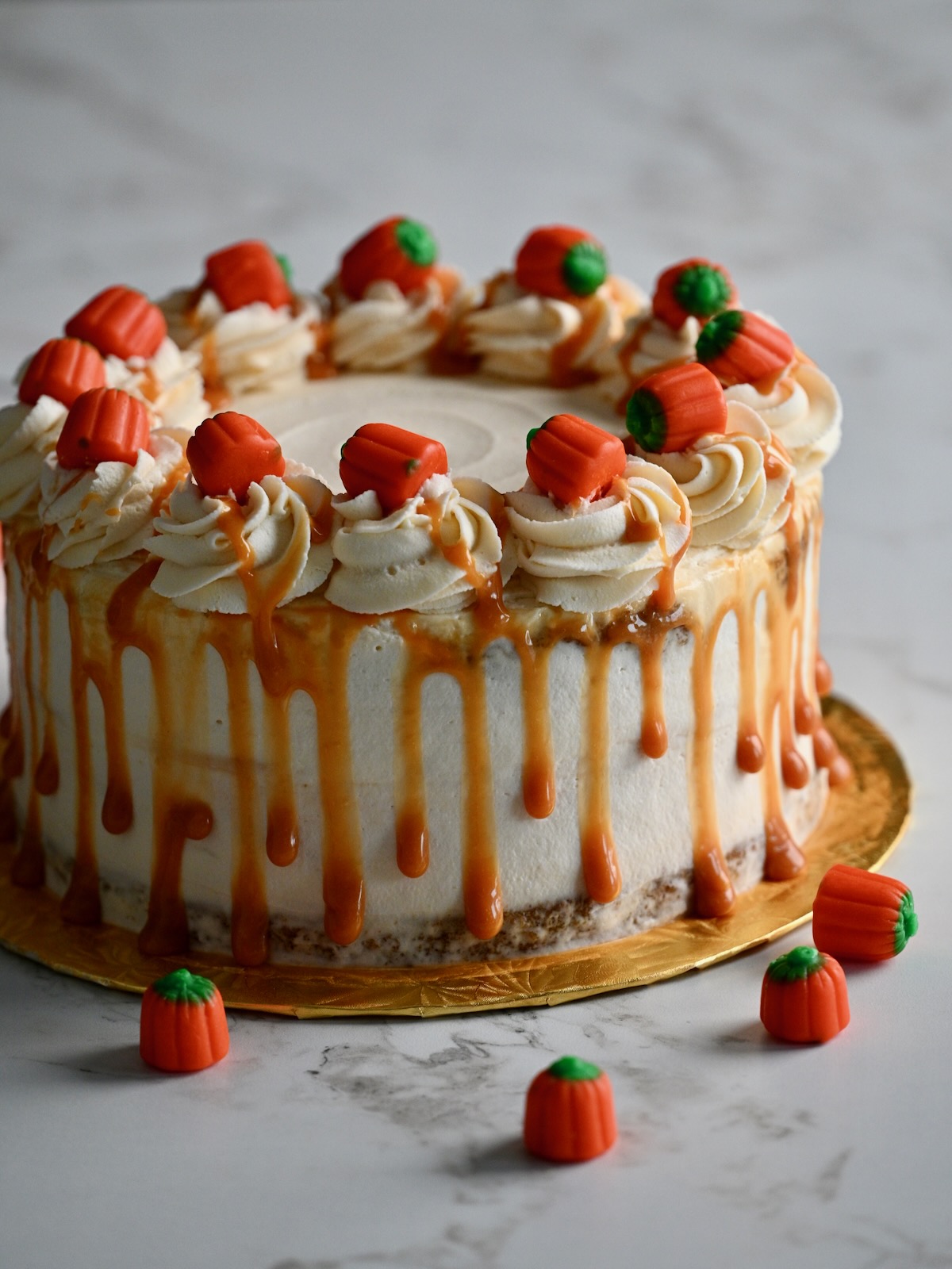 A whole pumpkin cake cheesecake topped with whipped cream swirls, candy pumpkins, and caramel drip on a gold cake board.
