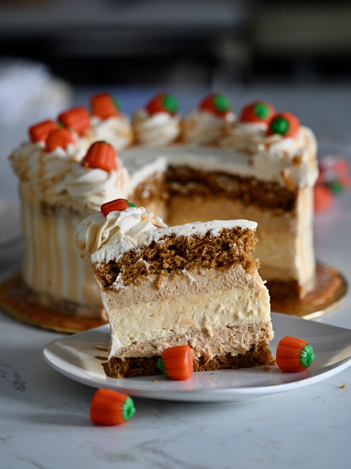 A tall slice of pumpkin cheesecake cake on a plate, topped with whipped cream swirls, candy pumpkins, and caramel drip.