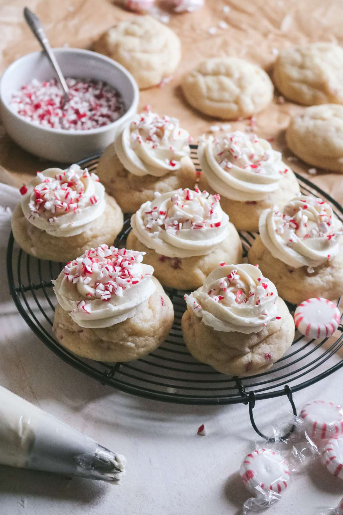 frosted peppermint sugar cookies on a wire rack with extra unfrosted cookies in the back and a bowl of crushed mint candies.