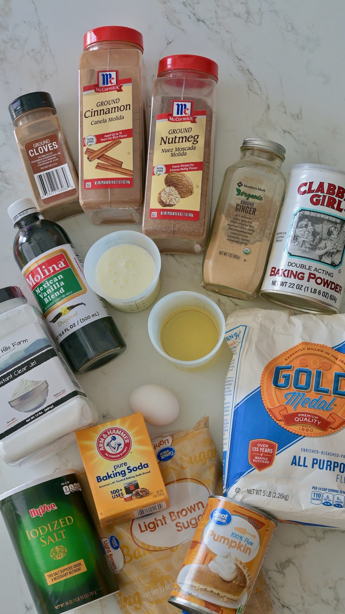Ingredients for the pumpkin cake layers displayed on a marble surface, including flour, brown sugar, pumpkin purée, baking powder, baking soda, cinnamon, nutmeg, ginger, cloves, vanilla, buttermilk, and vegetable oil.