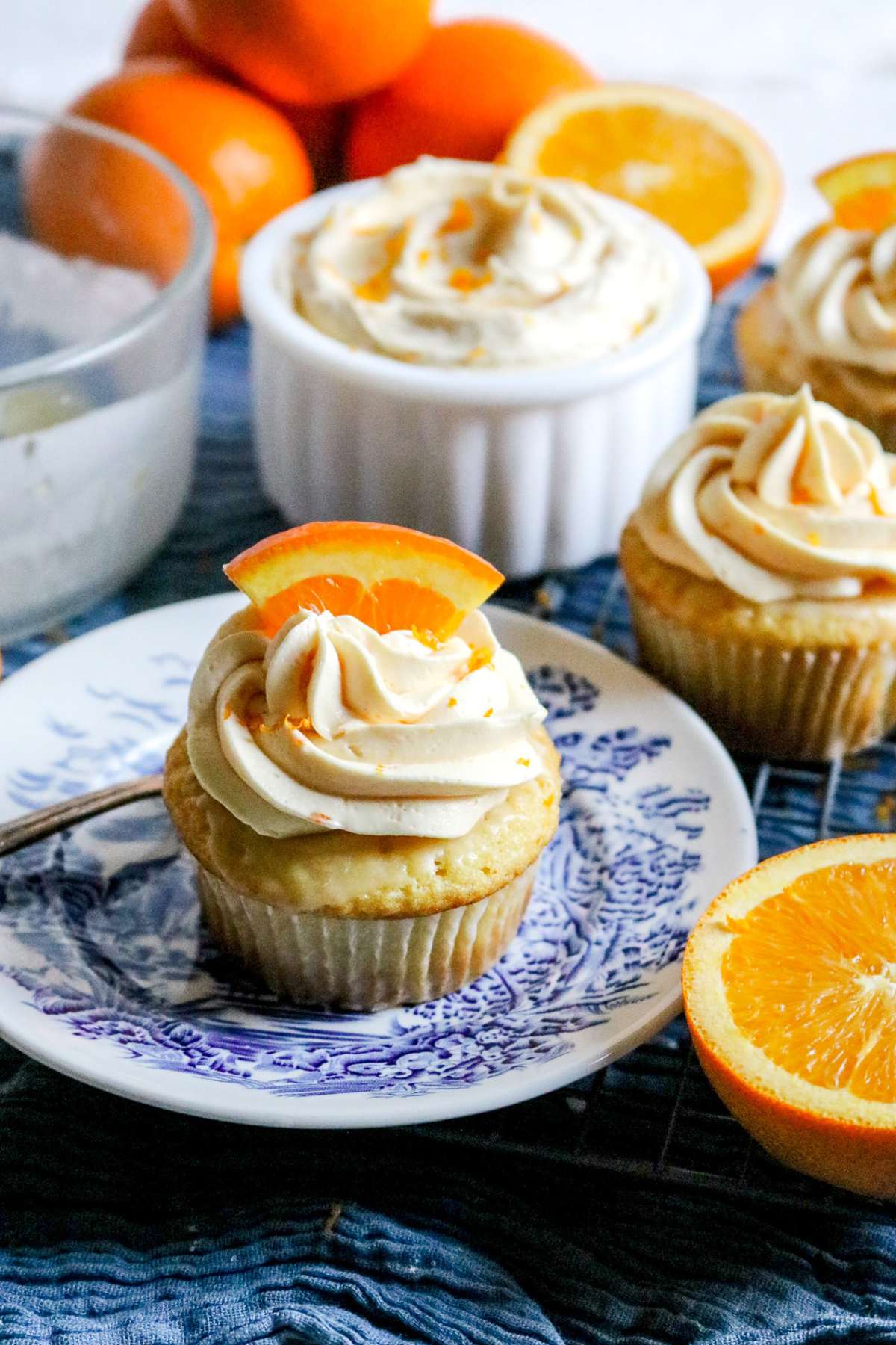 a scene with frosted orange cupcakes with a bowl of homemade buttercream, sliced and whole oranges, and more cupcakes.
