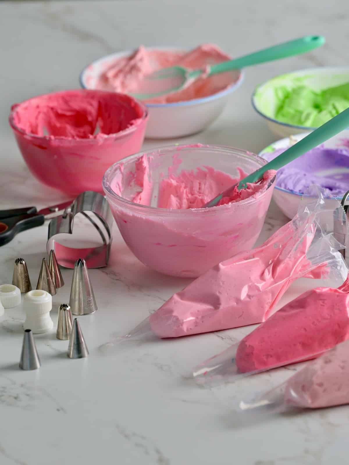 3 piping bags filled with light pink, pink, and magenta buttercream, with various bowls of buttercream frosting and piping tips and a heart cookie cutter in view on the table.