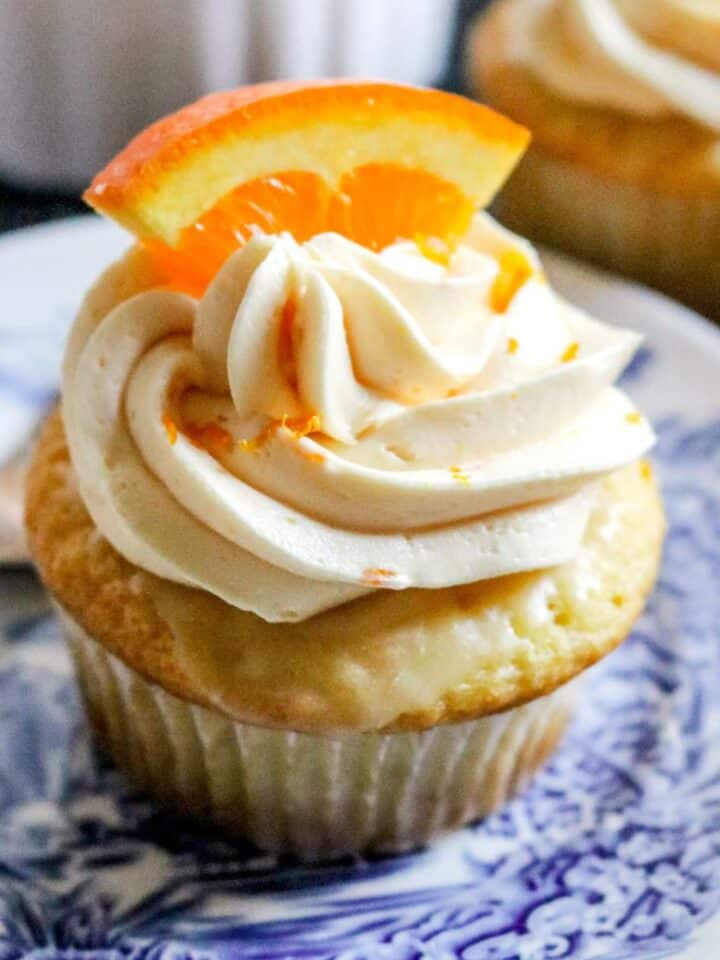 close up view of a fresh orange cupcake on a blue and white plate with orange buttercream and a sliced orange on top.