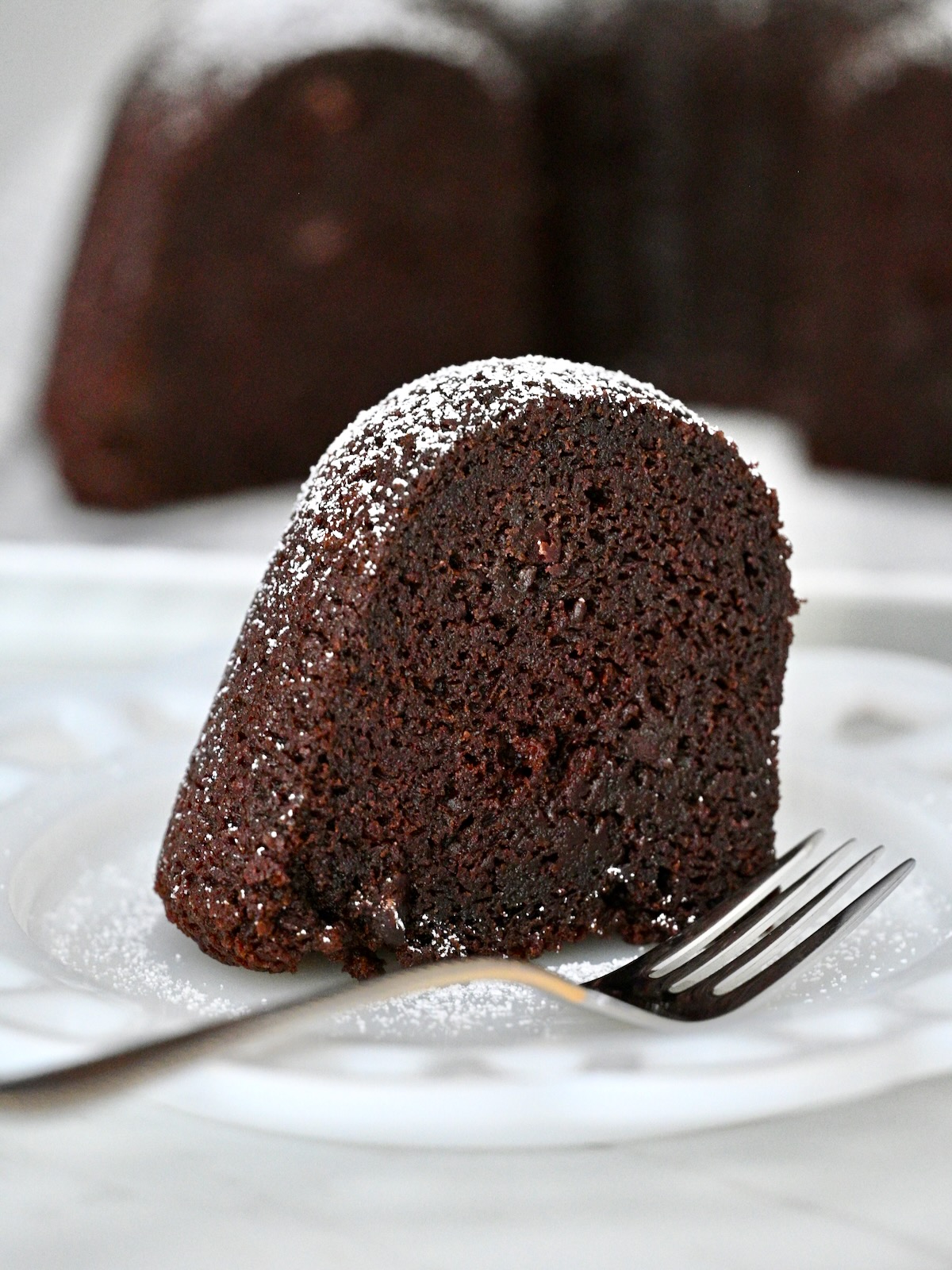 Single slice of moist chocolate bundt cake with chocolate chips and powdered sugar on a plate with a fork.