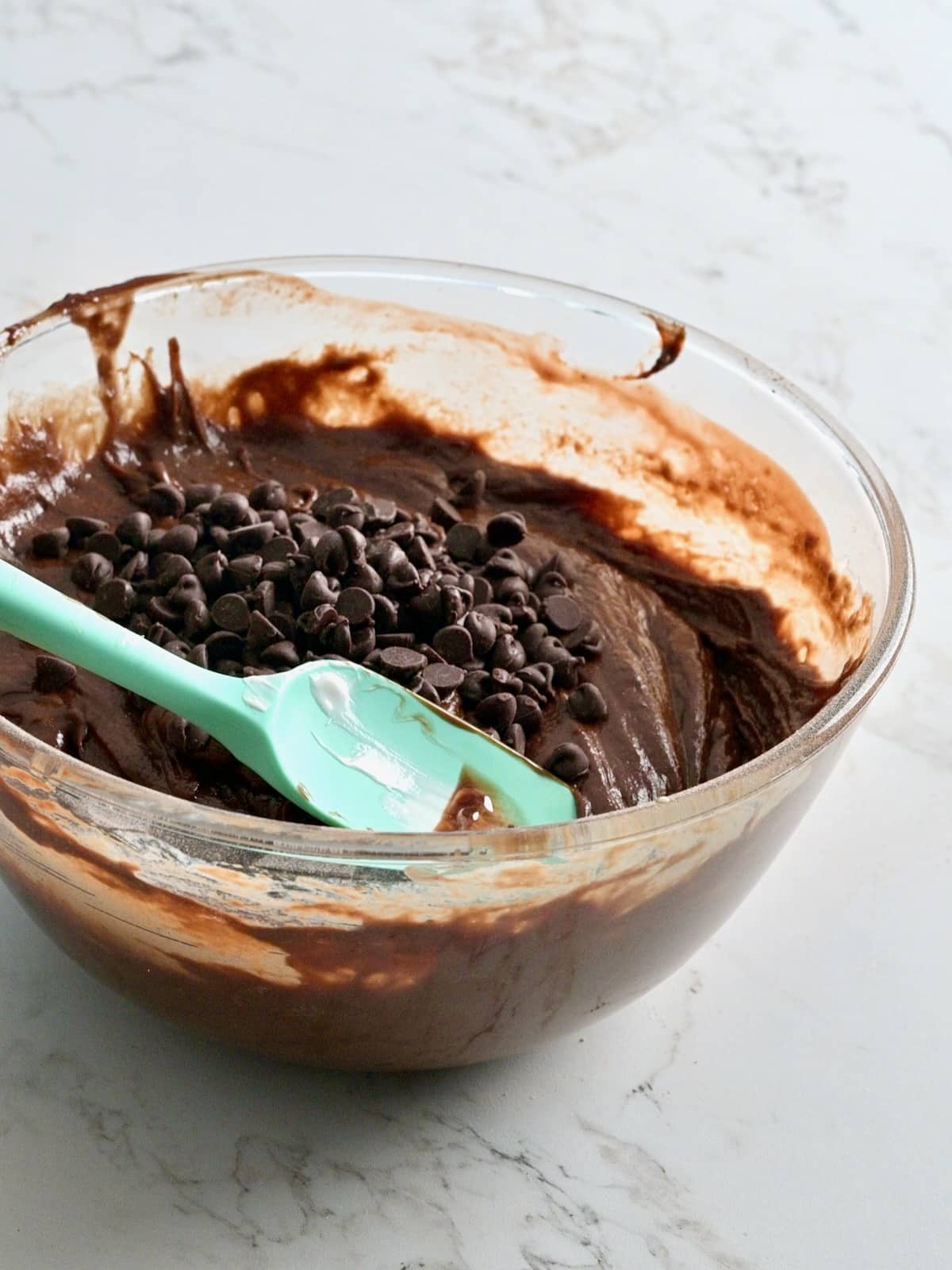 Glass bowl of thick chocolate bundt cake batter topped with semi-sweet chocolate chips and a teal spatula.