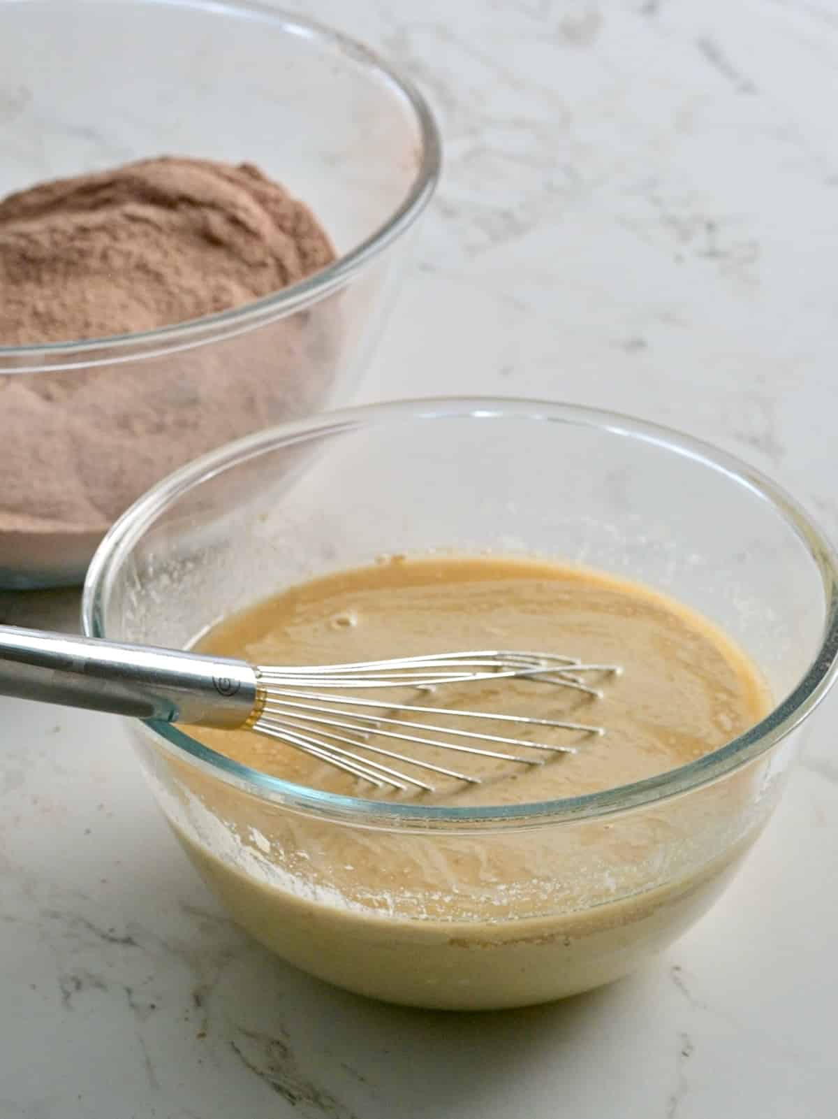 Whisked wet ingredients in a glass bowl next to a bowl of sifted dry ingredients for chocolate bundt cake.