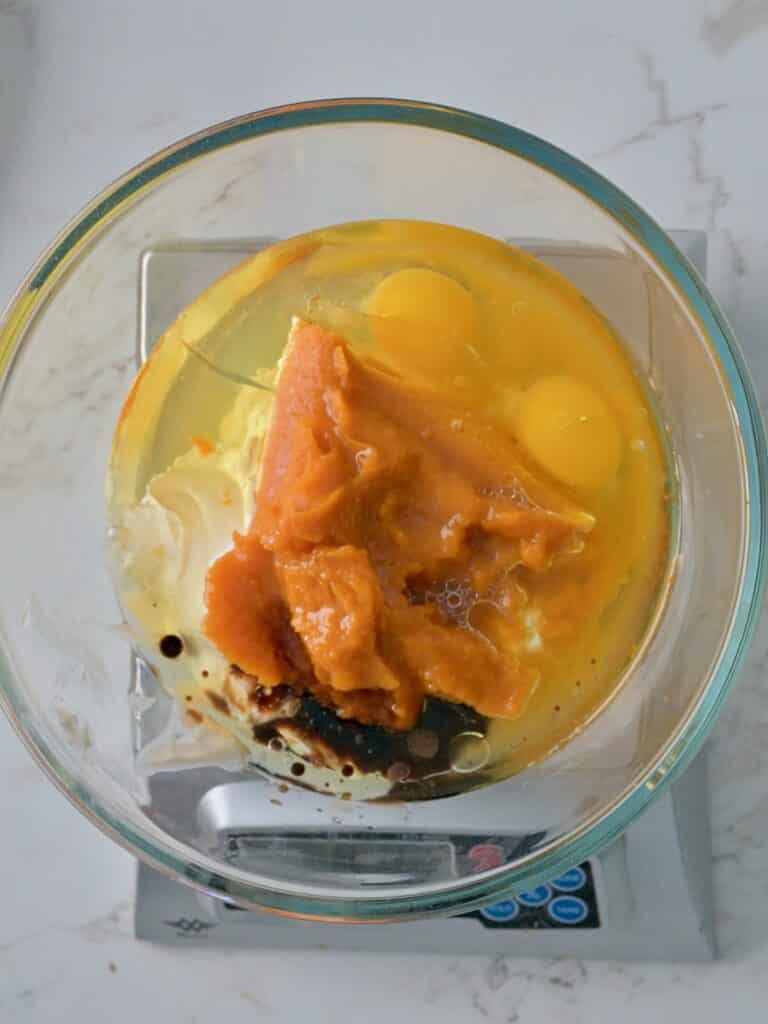 Pumpkin puree, eggs, and wet ingredients in a glass mixing bowl on a kitchen scale for pumpkin mini bundt cakes.