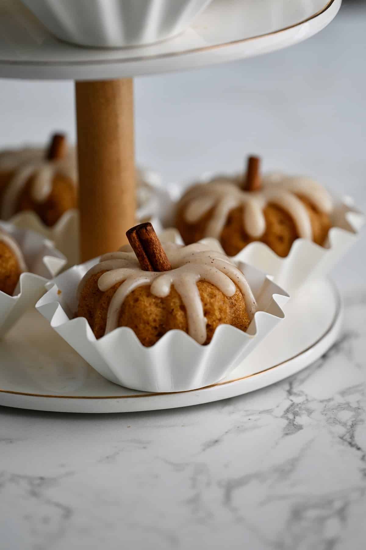 Pumpkin mini bundt cakes with cinnamon cream cheese glaze on the lower tier of a white dessert stand.