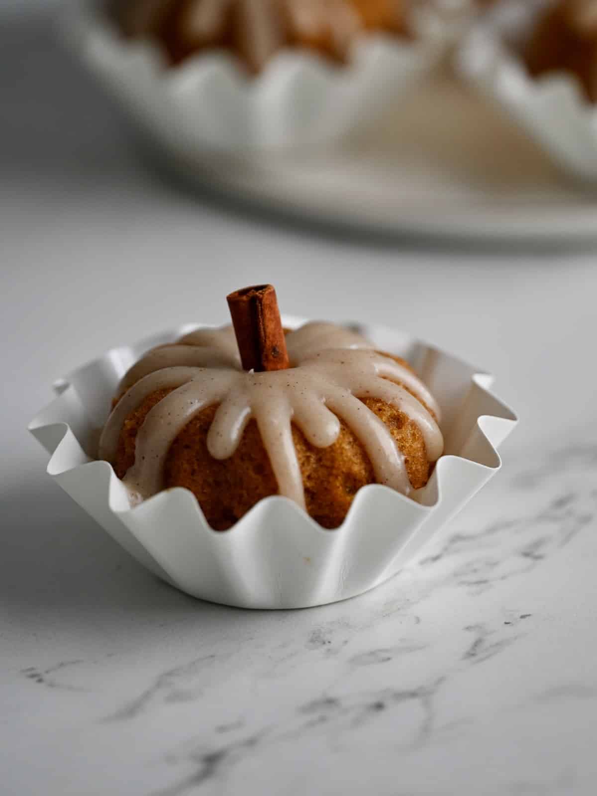 A single pumpkin mini bundt cake in a white fluted paper cup with cinnamon cream cheese glaze and a cinnamon stick stem.