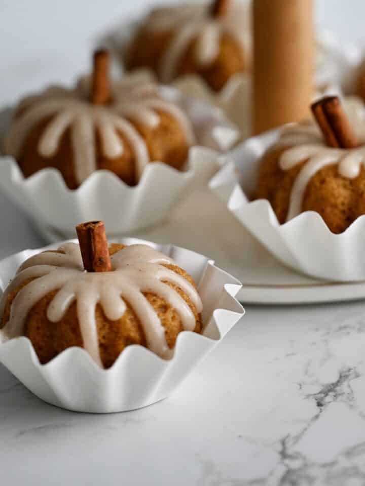 Glazed pumpkin mini bundt cakes in white fluted baking cups on a marble counter.